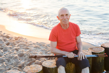 Old man in orange shirt and black shorts sitting on the beach with nordic walking poles. Senior man having rest on sea.