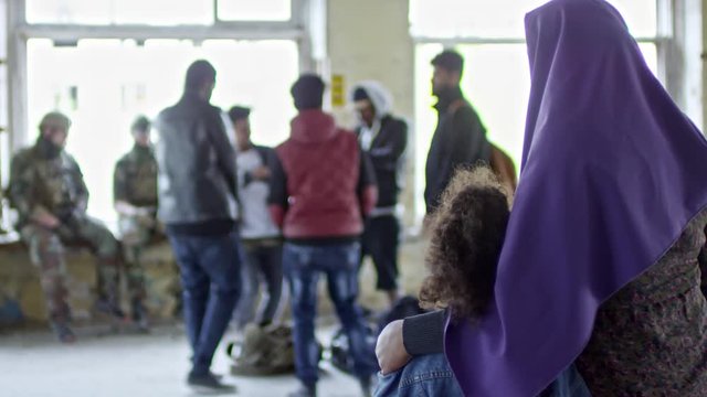 Rack focus of unrecognizable Muslim woman in purple niqab hugging little girl and looking at happy Arab male refugees dancing in destroyed building; Caucasian soldiers in uniform watching and chatting