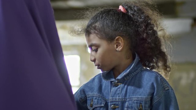 Close up of playful little Arab girl laughing and touching purple niqab of her unrecognizable mother; soldiers in uniforms walking in background