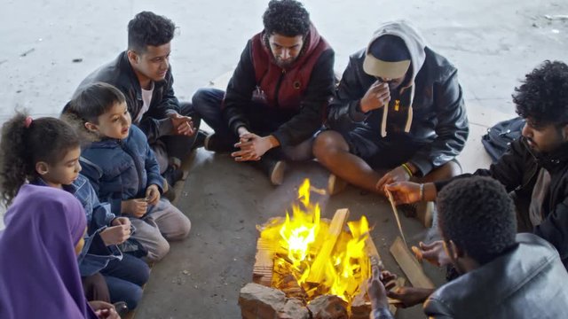 PAN with top view of Syrian men in jackets and little children with their Muslim mother in purple niqab sitting by fire in abandoned building