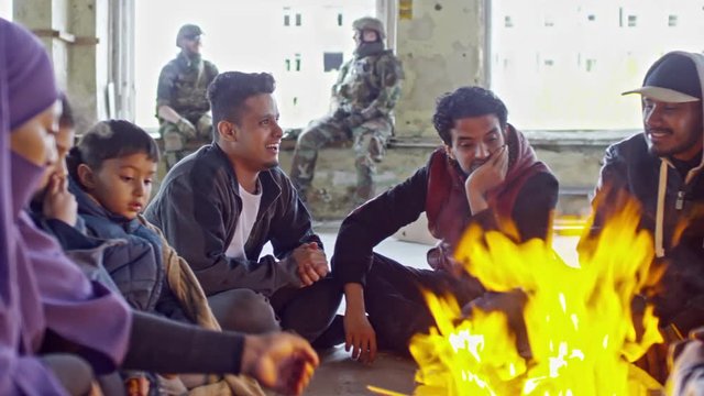 PAN of tired Arab male refugee and Muslim woman with little children wearing purple niqab warming their hands by fire in abandoned building; soldiers in uniforms chatting in background