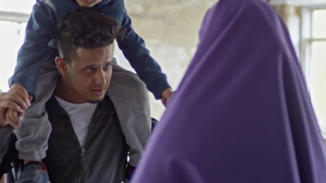 Tilt up of cheerful young Arab man holding little boy on shoulders and chatting with unrecognizable Muslim woman in purple niqab