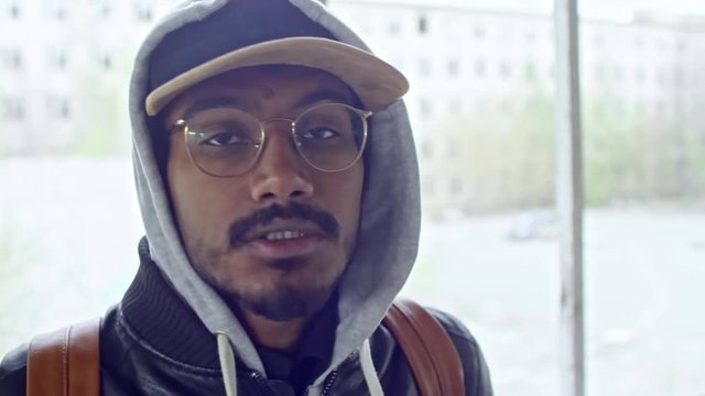 Close up of serious bearded Arab man in glasses wearing hood and jacket looking at camera and talking angrily, then pushing away camera and covering lens with hand