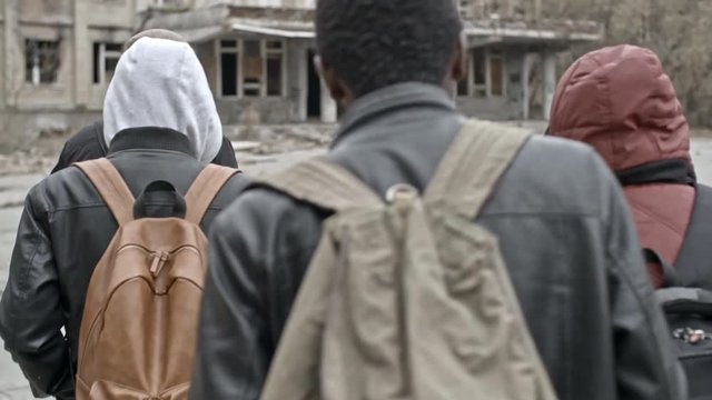 Tracking with rear view of group of Arab men with backpacks and Muslim woman in purple niqab walking towards abandoned building; soldiers in uniform patrolling