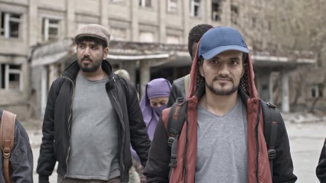 Dolly of group of serious male Arab refugees and Muslim woman in purple niqab walking towards camera; soldiers in uniforms patrolling before abandoned building in background