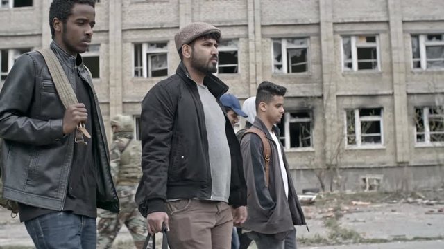 Tracking with side view of male Arab refugees wearing jackets and carrying backpacks walking before abandoned building in camp; soldiers in uniform and Muslim woman in purple niqab in background