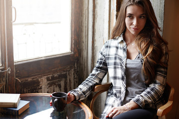 Fototapeta premium Casually dressed cute girl enjoying fresh cappuccino at coffee house. Confident young female having breakfast at shabby cafe, sitting by window with mug of hot drink, copy space for your information