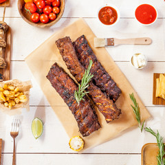 Spicy hot grilled spare ribs from a summer BBQ served with chips, corn and fresh tomatoes on an old wooden cutting board, top view