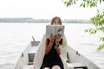 Obraz premium Portrait of an attractive woman wearing black polka dots dress, white shawl and glasses reading a book in a boat on a lake.