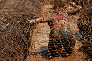 Mud race runners.Woman covered with mud fighting to get out of a net in the obstacle race - obrazy, fototapety, plakaty