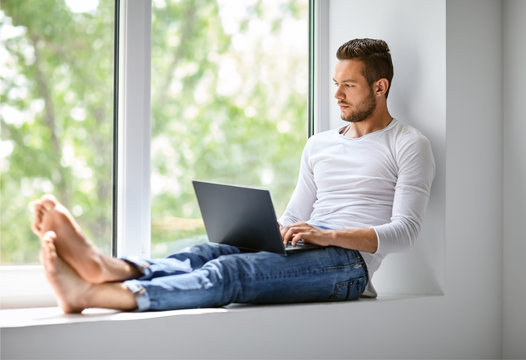 Handsome Man Typing On Laptop Sitting On Window Sill