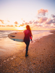 Fototapeta na wymiar Surf girl with surfboard on a beach at colorful sunset or sunrise.