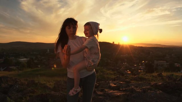 A happy family in the rays of the setting summer sun, mother is holding her daughter in her arms. Young woman and little girl make inviting gesture together.