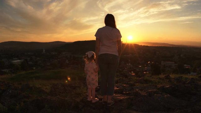 A happy family in the rays of the setting summer sun, mom holds her daughter by the hand, silhouettes of a woman and a little girl on a hill.