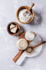 Rye and wheat sourdough in glass jars, fresh and instant yeast, olive wood bowl of flour for baking homemade bread. With spoon, serving board over gray concrete background. Top view