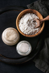 Rye and wheat sourdough in glass jars, olive wood bowl of flour for baking homemade bread. With scoop, serving board, textile over black burned wooden background. Top view