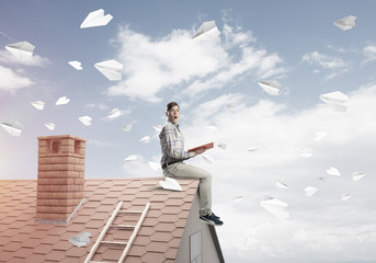 Man on brick roof reading book and paper planes flying in air