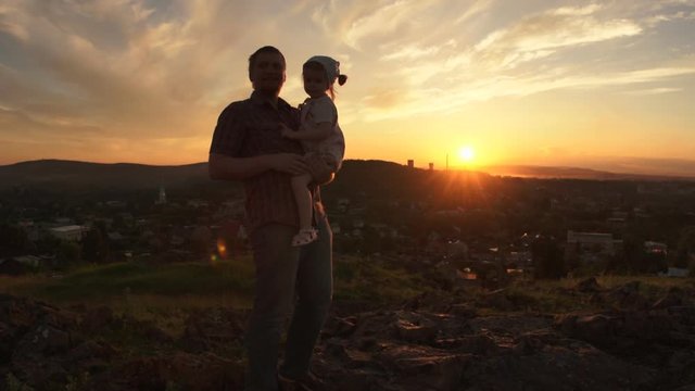 A happy family in the rays of the setting summer sun, the father holds his daughter in his arms, they have fun together. Silhouettes of a man and a little girl on a hill.
