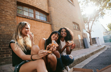 Beautiful girls sitting outdoors by the road eating pizza