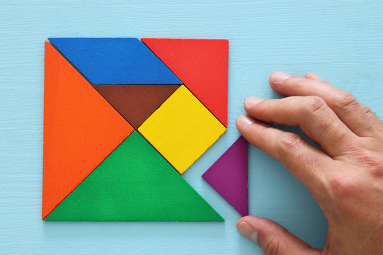man's hand holding a missing piece in a square tangram puzzle, over wooden table