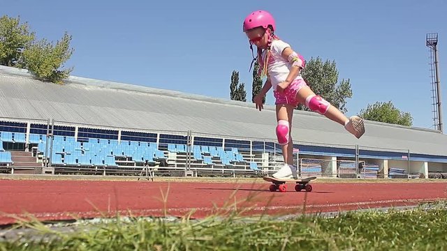 Pretty little girl learning to skateboard outdoors on beautiful summer day. Fashionably dressed girl, learn to skateboard on the stadium's treadmill.