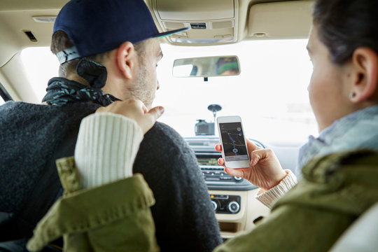 Young couple sitting in car, woman holding smartphone with map on screen, rear view