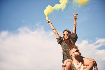 Young boho woman holding yellow smoke flare on boyfriend's shoulders at festival