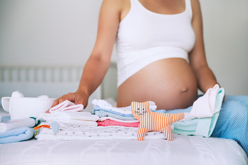 Pile of baby clothes, stuff and pregnant woman in home interior