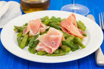 green beans with prosciutto on white plate on wooden background