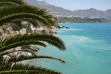 Fototapeta na wymiar The Spanish summer coast with people on the beach in the blue water and the mountain in the background and a palm tree in the foreground