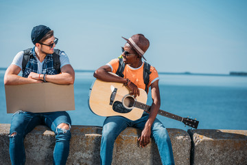 multicultural young men resting on parapet while hitchhiking during trip