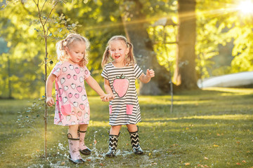 Naklejka premium The cute little blond girls in rubber boots playing with water splashes on the field in summer