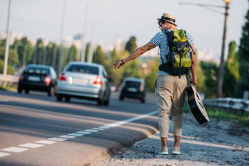 back view of young man with guitar gesturing to stop car while hitchhiking alone
