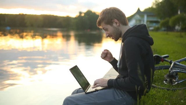Man freelancer working on the lake with a tablet computer, image at sunset. The lights of a sun. Gets good news. Experiencing Delight. Very happy