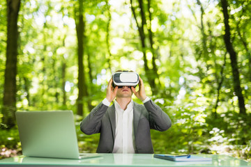 Confident handsome business man with virtual reality glasses pointed with hands at office desk in green park. Business concept.