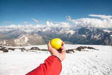 A man's hand holds an apple against the background of snow-capped mountains and snow underfoot.
