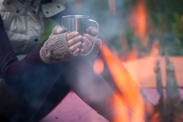 Hands in mitts holding hot tea cup outdoor near bonfire