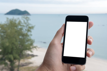 Mockup image of a hand holding black mobile phone with blank white screen with blue sky , white beach and the sea in background