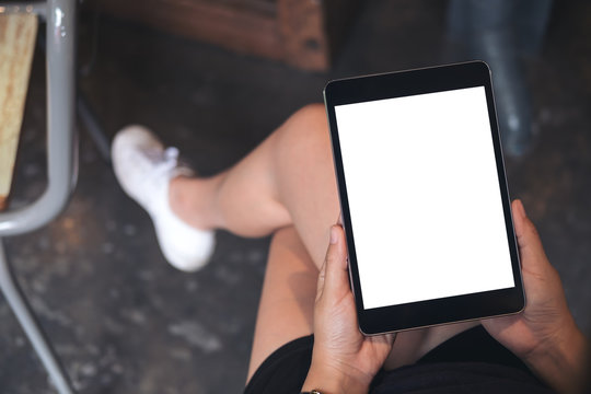 Mockup image of woman's hands holding black tablet pc with blank white screen on thigh with concrete floor background in modern cafe