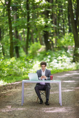 Obraz premium Businessman sitting at the office desk work at laptop computer and cup of coffee typing mobile phone on road of green forest park. Business concept.
