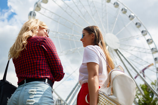 Two friendly girls standing by ferry in amusement park and deciding where to go