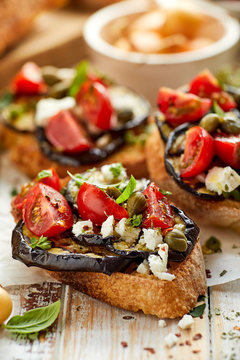 Bruschetta with grilled aubergine, cherry tomatoes, feta cheese, capers and fresh aromatic herbs, on a wooden table. Delicious Mediterranean appetizer