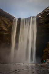 Obraz premium person standing admiring a huge waterfall at the side of a mountain with snow and ice in Iceland