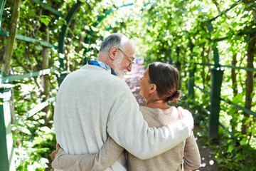 Happy embracing seniors walking along tree alley on sunny day