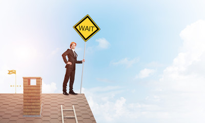 Young businessman on house brick roof holding yellow signboard. Mixed media