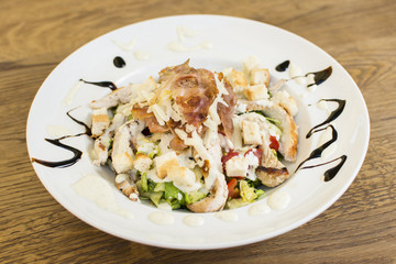 Healthy grilled chicken Caesar salad with cheese and croutons. Bacon and lettuce slices in a white plate on a wooden table.