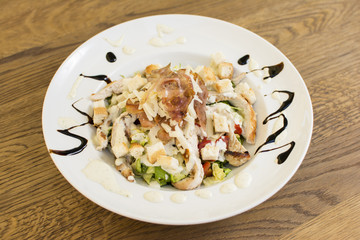 Healthy grilled chicken Caesar salad with cheese and croutons. Bacon and lettuce slices in a white plate on a wooden table.