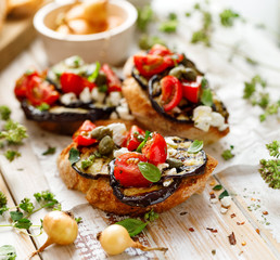 Bruschetta with grilled aubergine, cherry tomatoes, feta cheese, capers and fresh aromatic herbs, on a wooden table. Delicious Mediterranean appetizer - obrazy, fototapety, plakaty