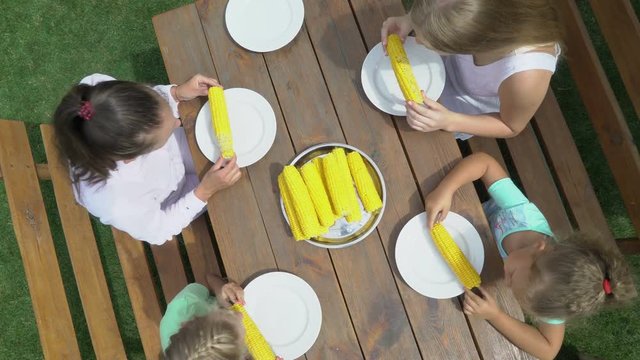 Family corn eating contest in the garden. Children have fun eats sweet corn cob at a wooden table outside. Top view of eaters on green grass lawn.