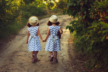 Happy twin sisters children. Girls sister in a park , walking on the road, holding hands. Sunlight and view from back.